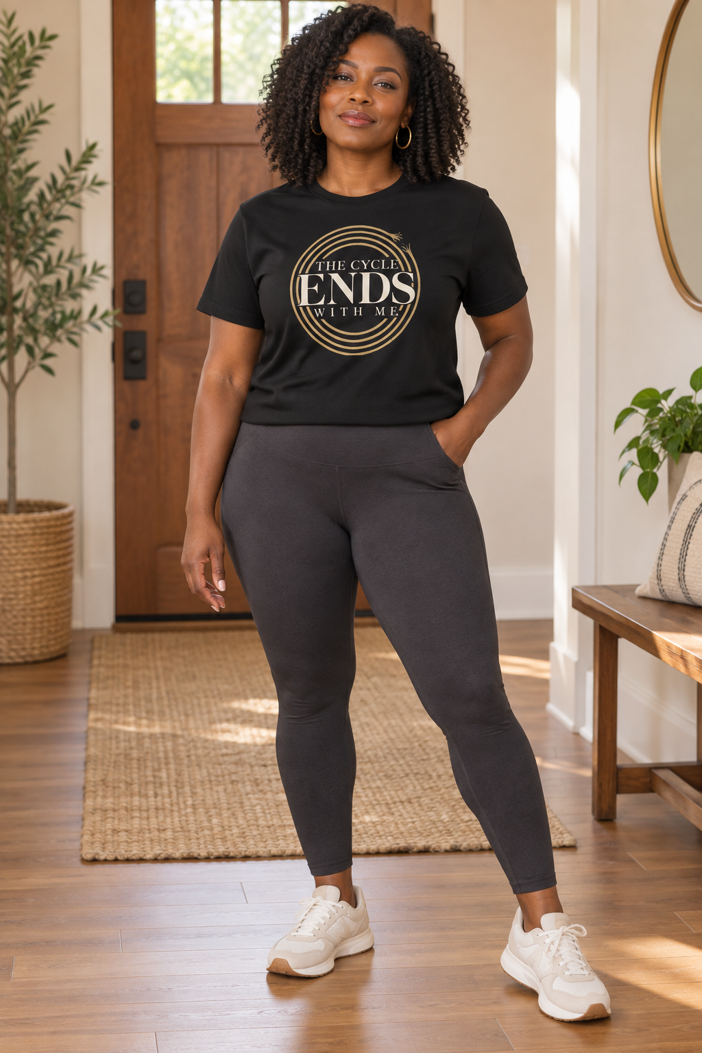 Black woman wearing The Cycle Ends With Me black tee with charcoal leggings in a warm, neutral home.