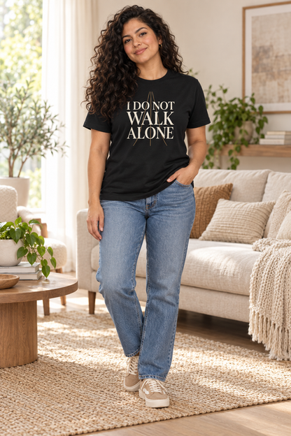 Latina woman wearing the I Do Not Walk Alone black tee with relaxed blue jeans in a sunlit living room.