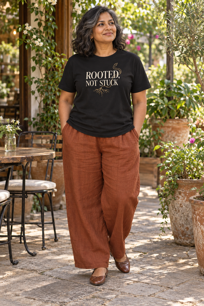 South Asian woman wearing the Rooted, Not Stuck black tee with rust linen trousers on a plant-filled patio.
