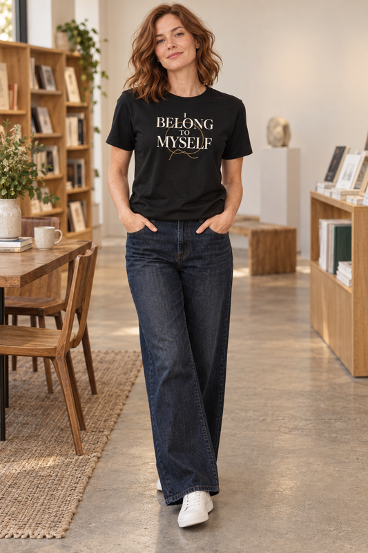 Red-haired woman wearing the I Belong to Myself black tee with dark-wash jeans in a warm bookstore setting.