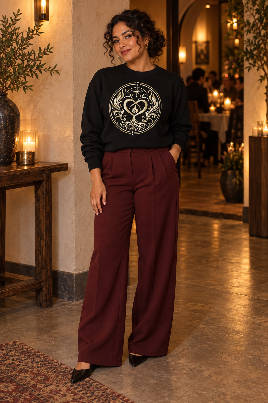 Woman wearing the black Sacred Connection Seal sweatshirt with tailored burgundy trousers in an elegant indoor setting.